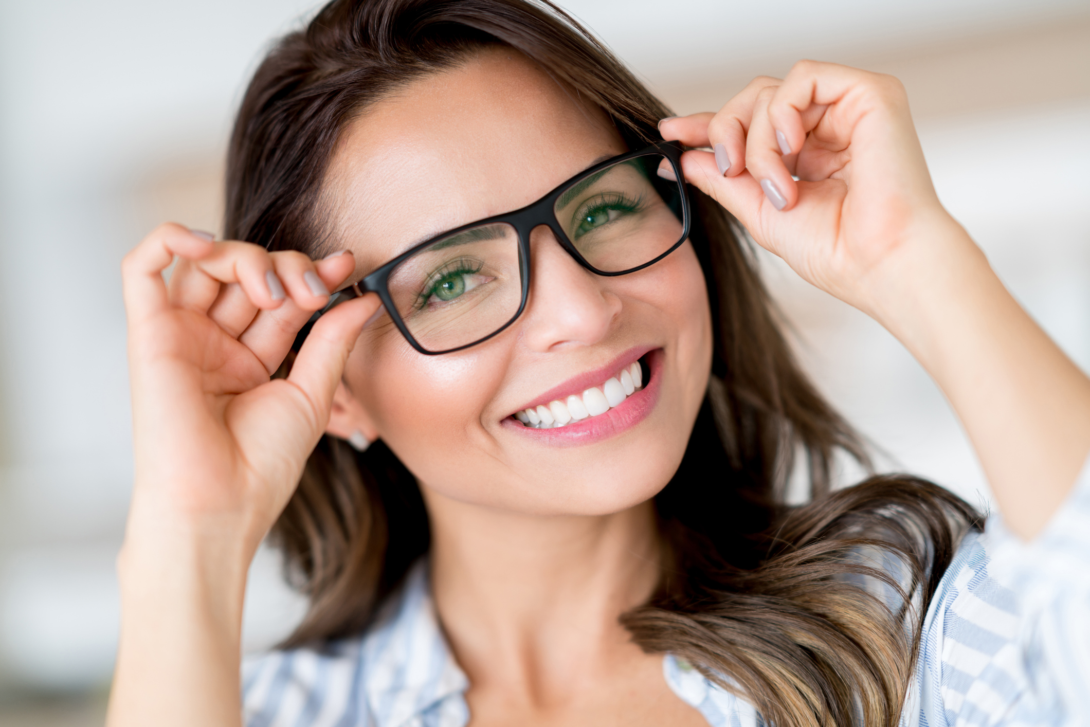 A smiling woman with long brown hair and green eyes adjusts her black-rimmed eyeglasses with both hands. She is wearing a plaid shirt and appears cheerful in a bright indoor setting. - Claye Eye Physicians & Surgeons - the best eye doctor in Fleming Island, Florida