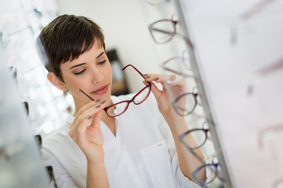 A person with short dark hair wearing a white shirt examines a pair of eyeglasses in an optical store, surrounded by displays of various frames. - Claye Eye Physicians & Surgeons - the best in Jacksonville, Florida